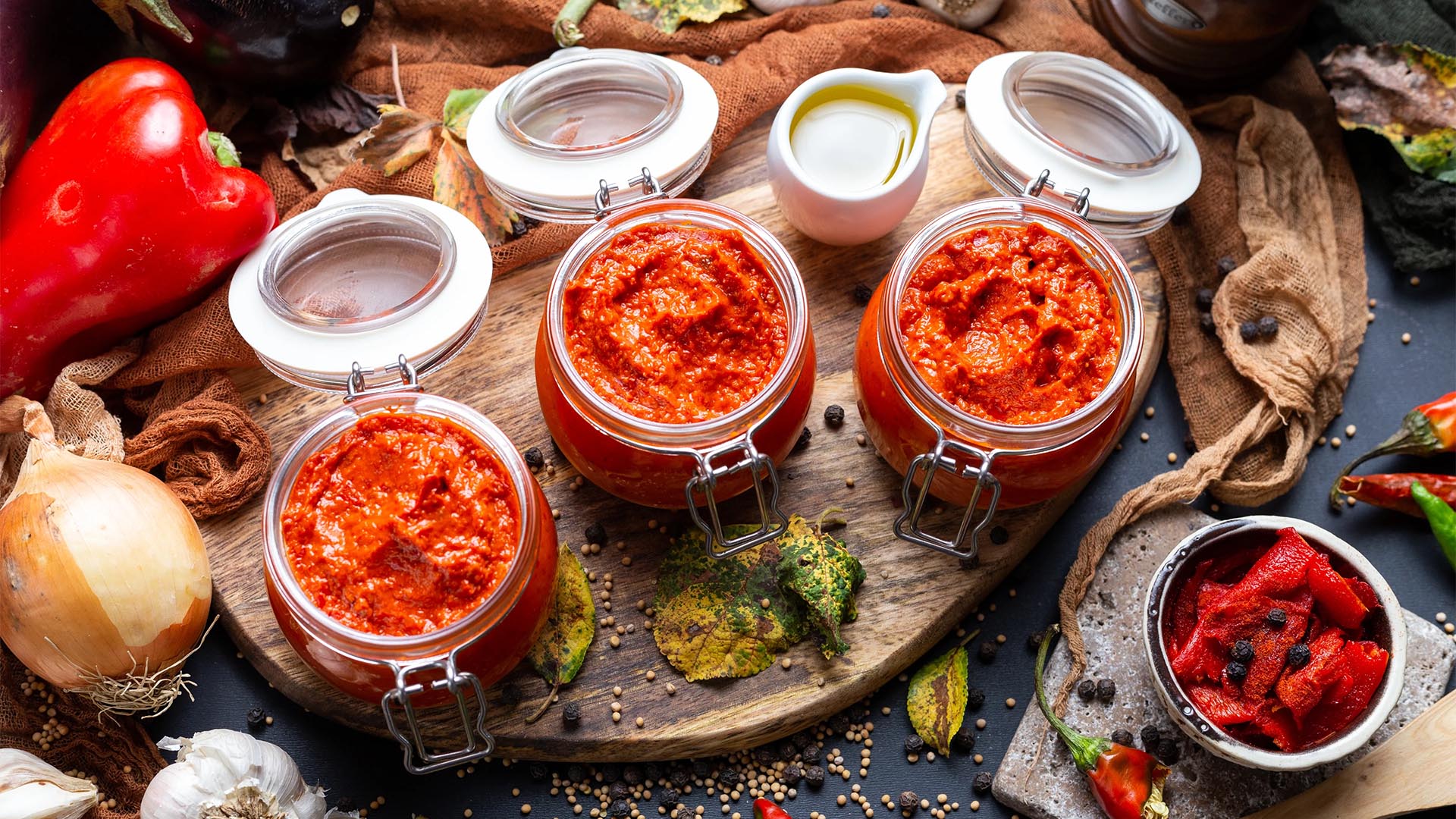 Jars of homemade relish surrounded by fresh ingredients including onions, garlic, and roasted peppers